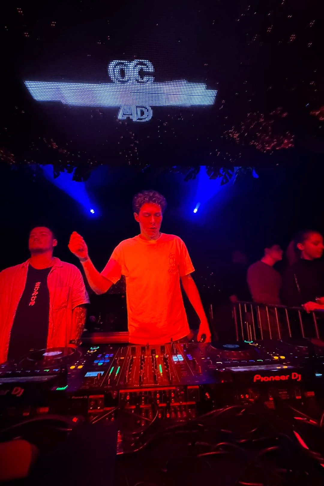 FØR U DJ in an orange T-shirt raising his hand behind Pioneer DJ decks under blue stage lights